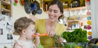 A lovely and natural snapshot of a woman and a toddler in a cozy kitchen. The woman is feeding the girl a big carrot. There are lots of fresh veggies on the counter.