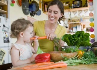 A lovely and natural snapshot of a woman and a toddler in a cozy kitchen. The woman is feeding the girl a big carrot. There are lots of fresh veggies on the counter.
