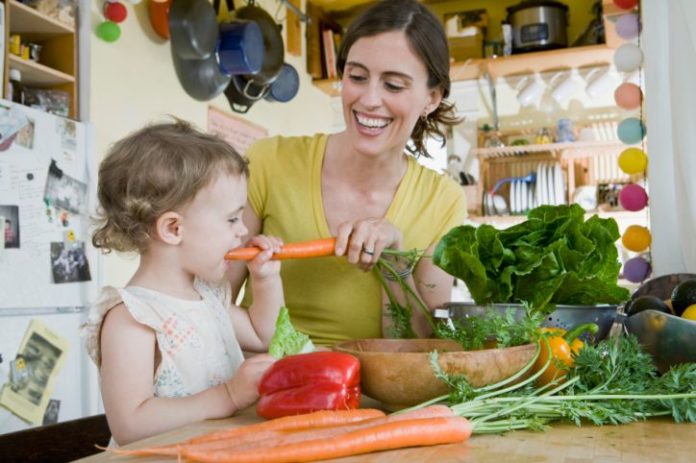 A lovely and natural snapshot of a woman and a toddler in a cozy kitchen. The woman is feeding the girl a big carrot. There are lots of fresh veggies on the counter.