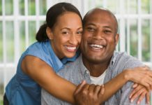 A couple in their late thirties or early forties smile to camera. The woman is hugging around the man's shoulders.