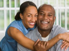 A couple in their late thirties or early forties smile to camera. The woman is hugging around the man's shoulders.