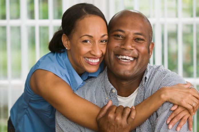 A couple in their late thirties or early forties smile to camera. The woman is hugging around the man's shoulders.