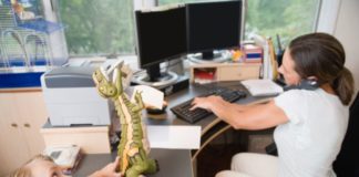 Mum works at her home office while little boy plays with a dinosaur toy.