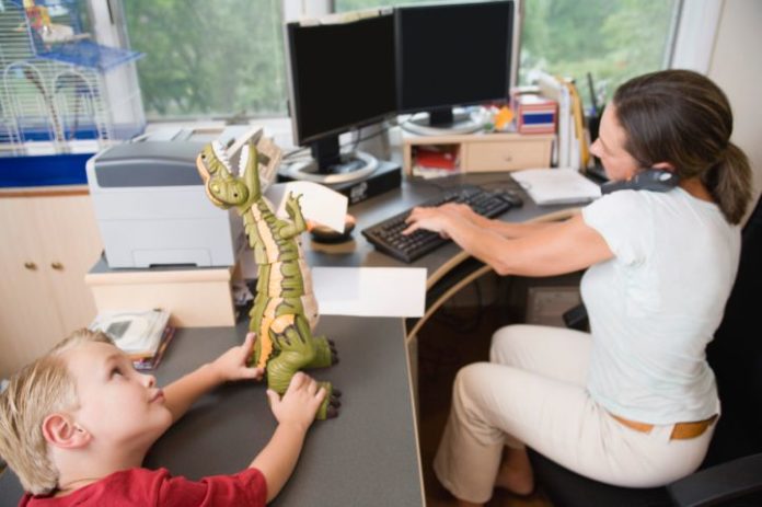 Mum works at her home office while little boy plays with a dinosaur toy.