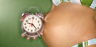 A very pregnant woman hold a clock in front of her belly.