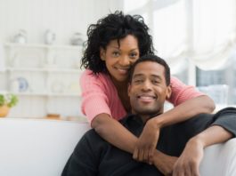 In a fresh looking white house, a man sits on a sofa while a woman hugs him around the neck.