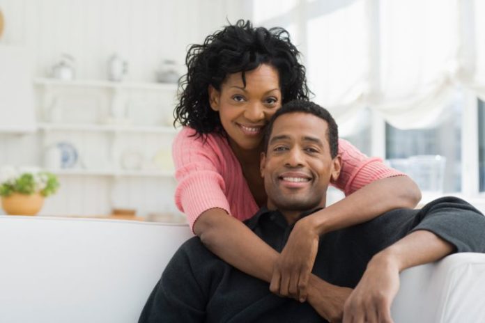 In a fresh looking white house, a man sits on a sofa while a woman hugs him around the neck.