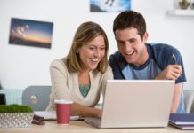 A happy college age couple are using a single generic laptop. They both smile at the screen.