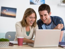 A happy college age couple are using a single generic laptop. They both smile at the screen.