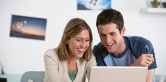A happy college age couple are using a single generic laptop. They both smile at the screen.