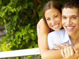 A very happy young couple with perfect teeth smile to the camera. They are outside. Perhaps on a park bench.