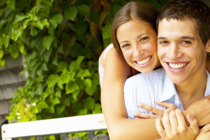 A very happy young couple with perfect teeth smile to the camera. They are outside. Perhaps on a park bench.