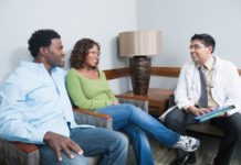 A couple consult with a doctor in what looks to be a waiting room. Everyone is smiling.