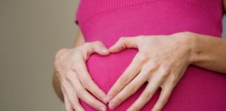 A woman holds her hands in the shape of a heart over her pregnant stomach.