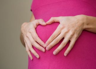 A woman holds her hands in the shape of a heart over her pregnant stomach.