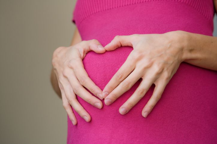 A woman holds her hands in the shape of a heart over her pregnant stomach.