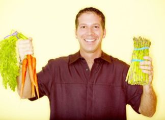 A smiling man in a burgundy button up holds carrots in one hand and asparagus in the other.