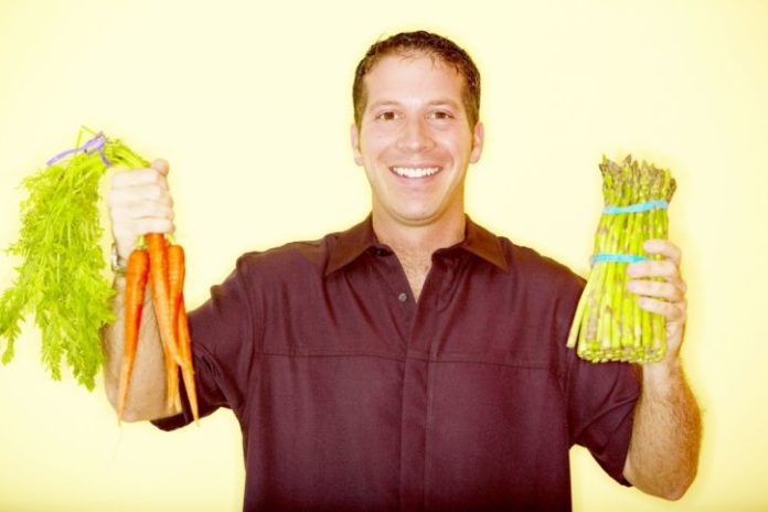 A smiling man in a burgundy button up holds carrots in one hand and asparagus in the other.
