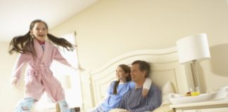 A girl jumps on the bed as her parents look on smiling.