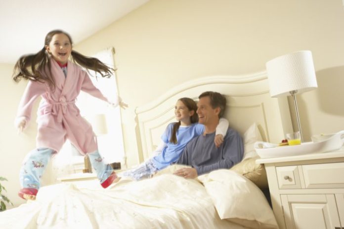 A girl jumps on the bed as her parents look on smiling.