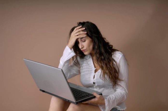 Woman sits with laptop on knees. She is distressed.