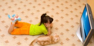 A girls lays on the floor watching TV with her cat.