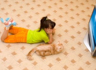 A girls lays on the floor watching TV with her cat.