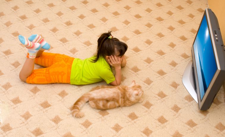 A girls lays on the floor watching TV with her cat.