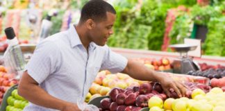 A man happily chooses a reddish apple from a produce shelf in a grocery store.