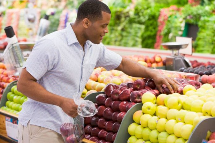 A man happily chooses a reddish apple from a produce shelf in a grocery store.