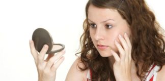 A teen girl touches her face while looking at herself in a compact mirror.