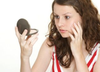 A teen girl touches her face while looking at herself in a compact mirror.