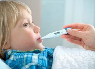 A hand holding a thermometer in a boy's mouth. The boy is in bed.