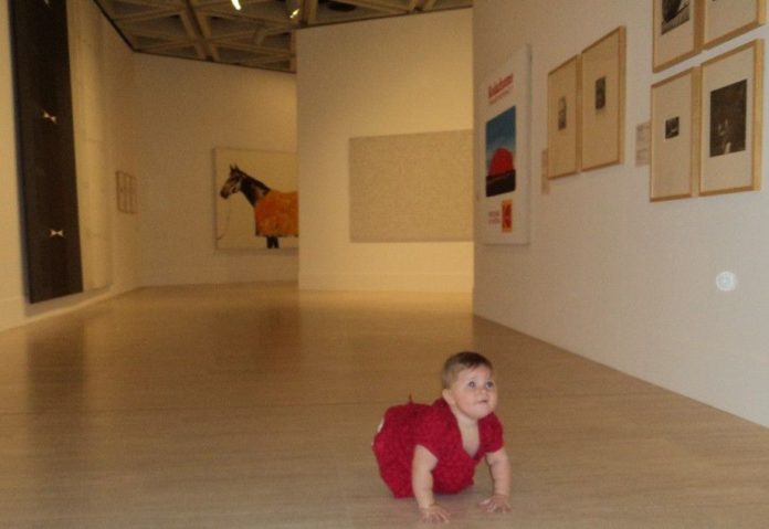 A little girl crawling on the floor of the art gallery, looks up to admire the paintings.