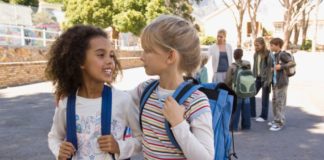 Two girls are in the school yard and both are wearing blue backpacks.