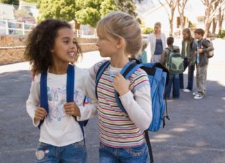 Two girls are in the school yard and both are wearing blue backpacks.