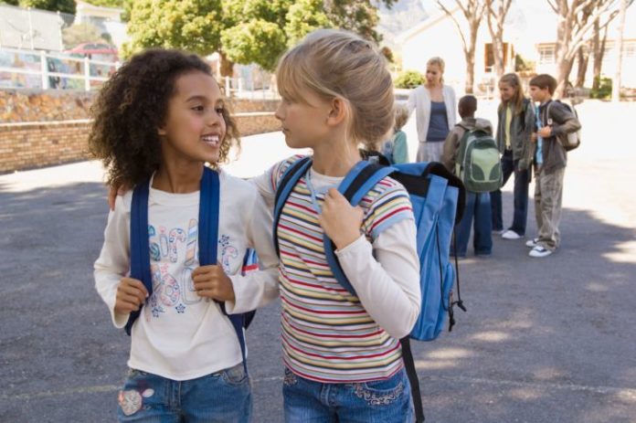 Two girls are in the school yard and both are wearing blue backpacks.